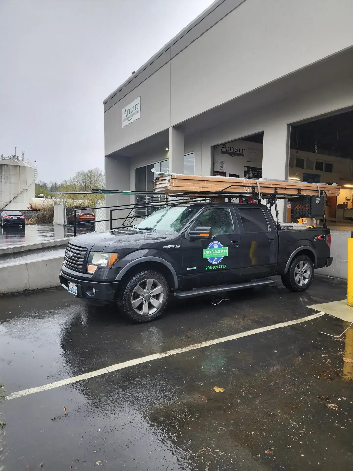 Black Ford F-150 truck with ladder rack and boards, Dan's Garage Door logo
