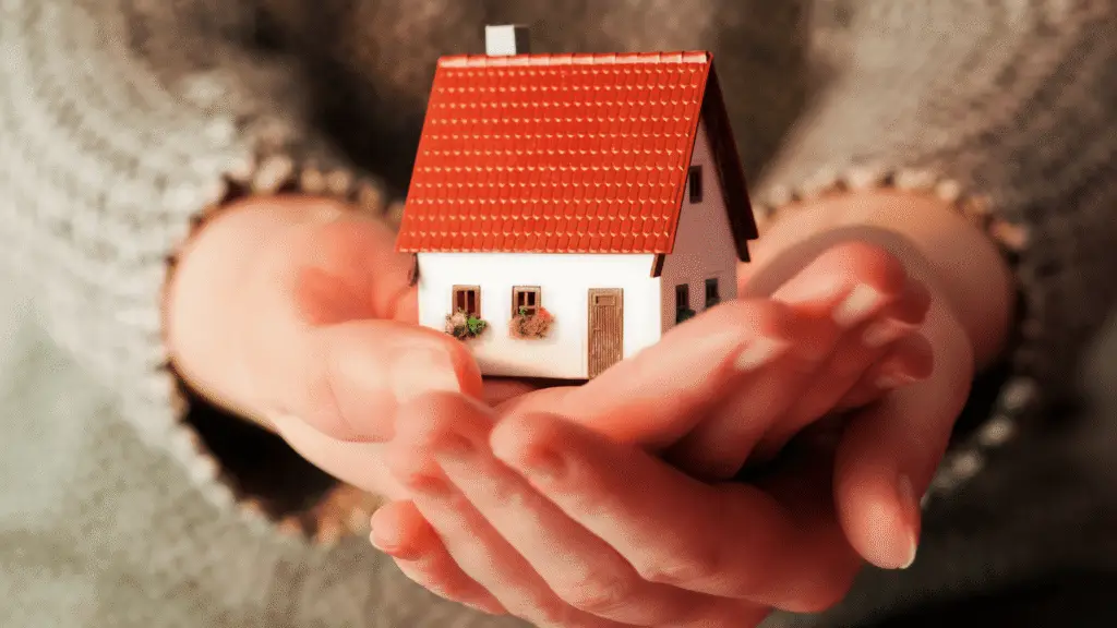 Hands holding a small model house with a red roof, symbolizing protection and care for a home.