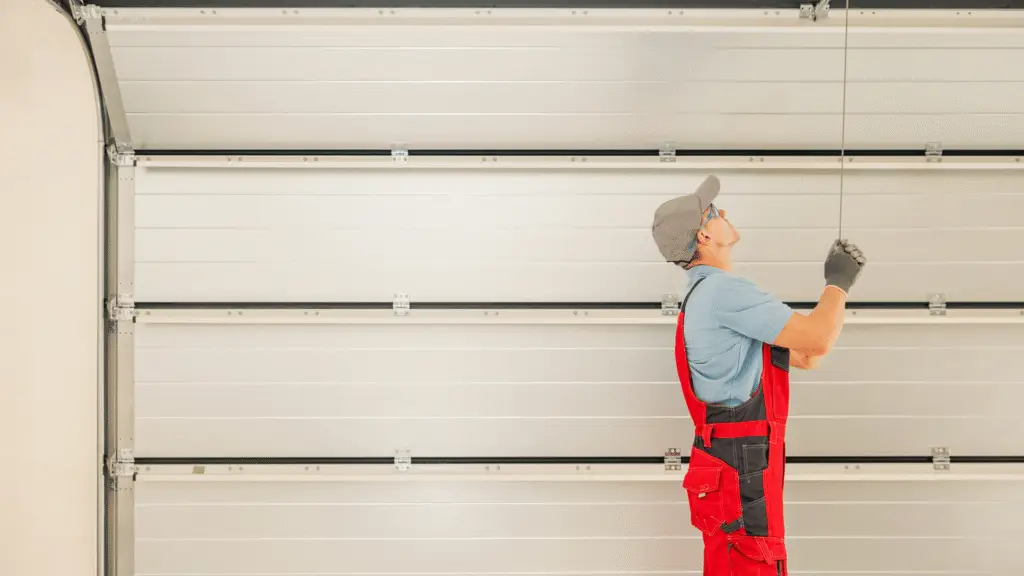 Worker in red overalls and a blue shirt installing or adjusting a garage door mechanism inside a garage.