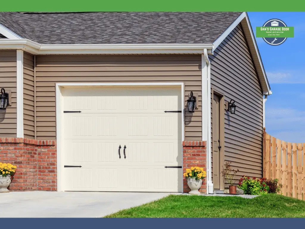 White garage door with black handles beside a man door, surrounded by flowers and a wooden fence.