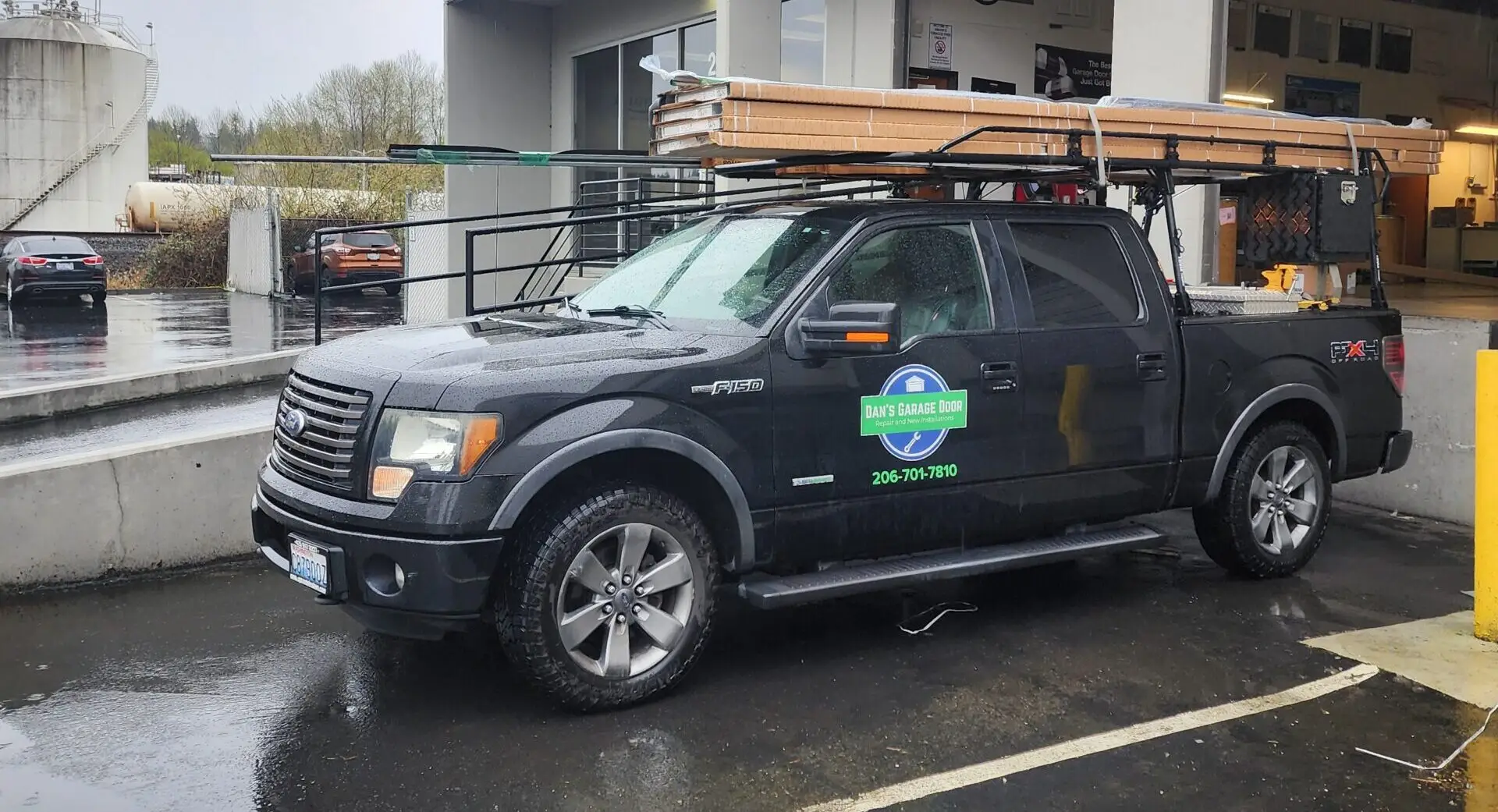 Black Ford F-150 pickup with ladder rack and lumber on top, wet lot