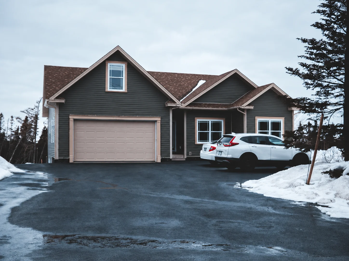 House with closed garage door, snowy driveway, and parked cars