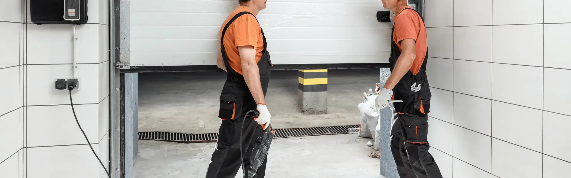 Two workers in orange shirts standing in a garage entrance, one holding a power tool